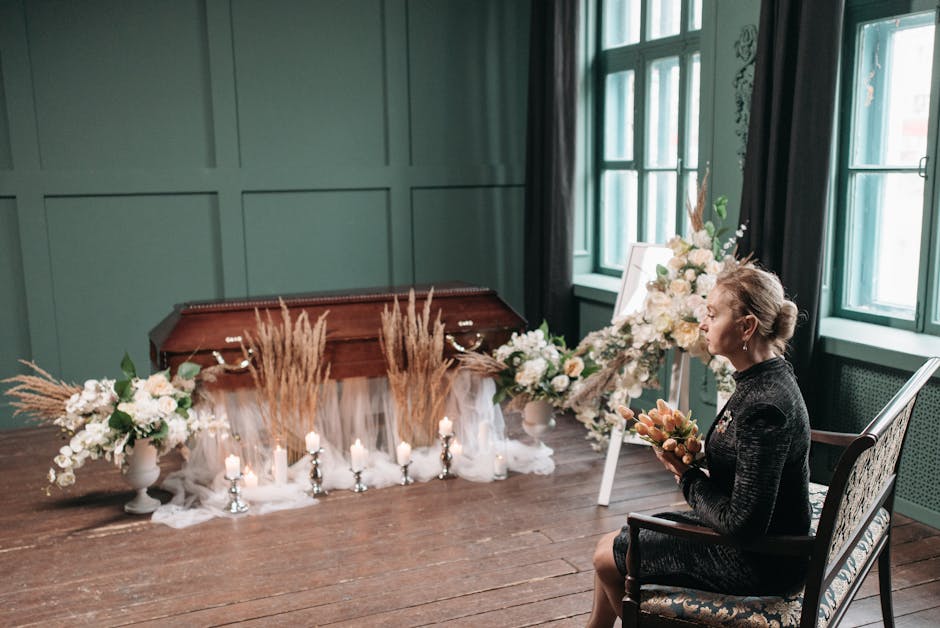 A woman seated on a decorative chair in a green-walled room holding a bouquet of soft pink and peach flowers. In front of her, there is a wooden table draped with sheer white fabric, decorated with la