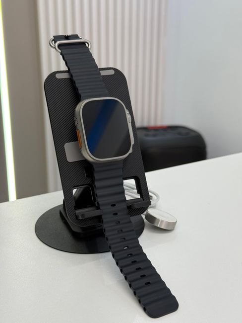A close-up of a smartwatch with a rectangular face and a blank screen, mounted on a black textured stand with a curved support and a cutout slot to hold the watch securely. The watch has a dark strap
