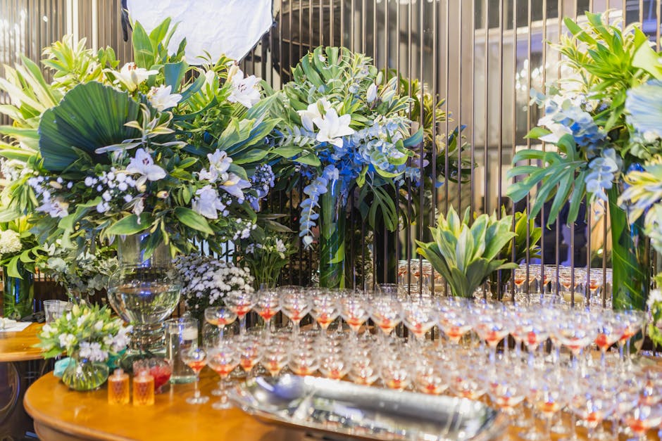 An elegant floral display featuring large arrangements of white lilies, blue delphiniums, and lush green foliage, including monstera and philodendron leaves, positioned behind a table set for a celebr