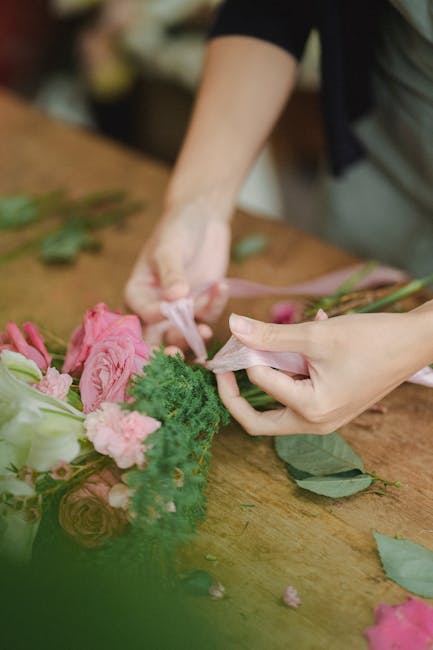 Close-up of a florist's hands assembling a floral arrangement on a wooden table, featuring pink roses, green foliage, and pink ribbon. The florist is wrapping the bouquet with light pink tissue paper,