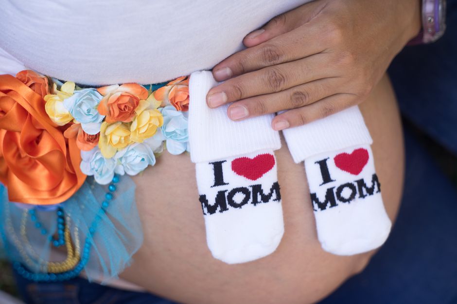 Close-up of a pregnant woman's abdomen decorated with a floral belt made of artificial roses in pastel shades of orange, yellow, and light blue, with a large orange satin ribbon bow on the side. She i