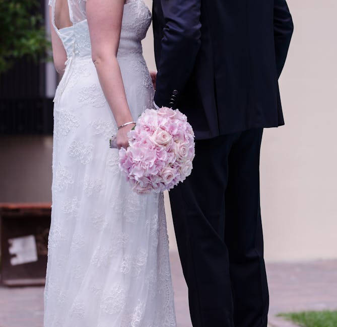 A close-up view of a bride and groom standing side by side during their wedding, with only the lower half of their bodies visible. The bride is wearing a white lace wedding gown with intricate floral 