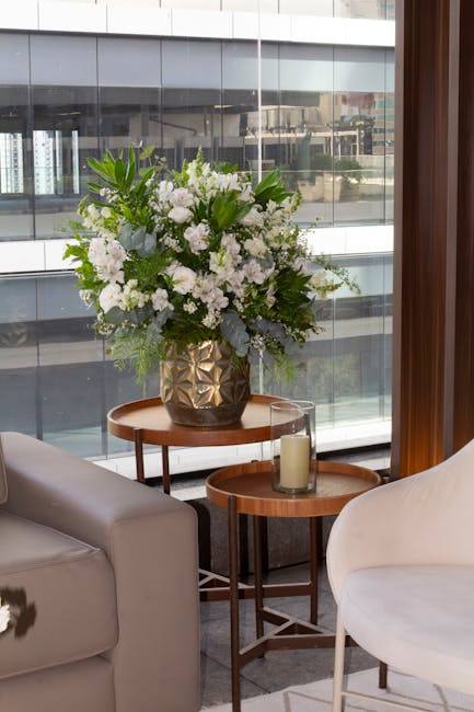 A tall floral arrangement of predominantly white flowers, including roses, hydrangeas, and lisianthus, is displayed in a dark, geometrically textured ceramic vase. The bouquet features lush green foli